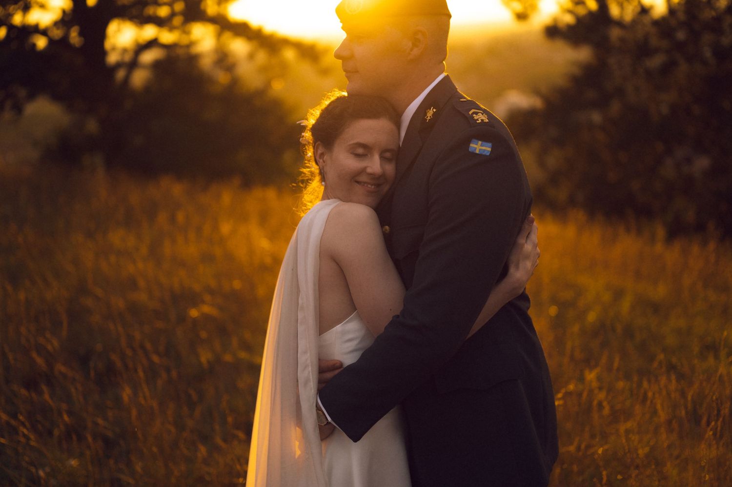 Bride and groom embracing at golden sunset in a field, glowing backlight creating romantic wedding portrait.
