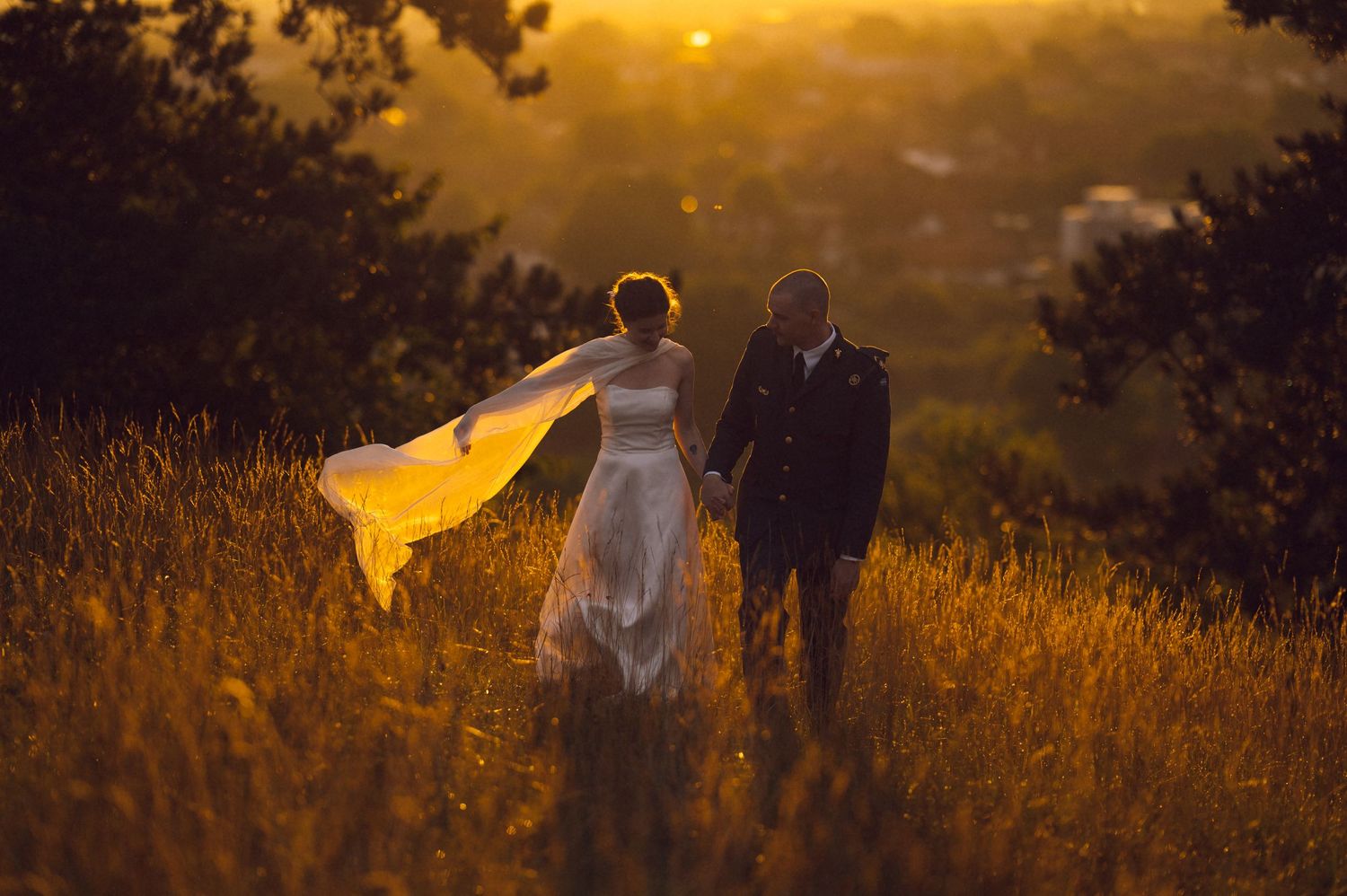 Bride and groom walking hand in hand through golden meadow at sunset, wedding veil flowing in the breeze.