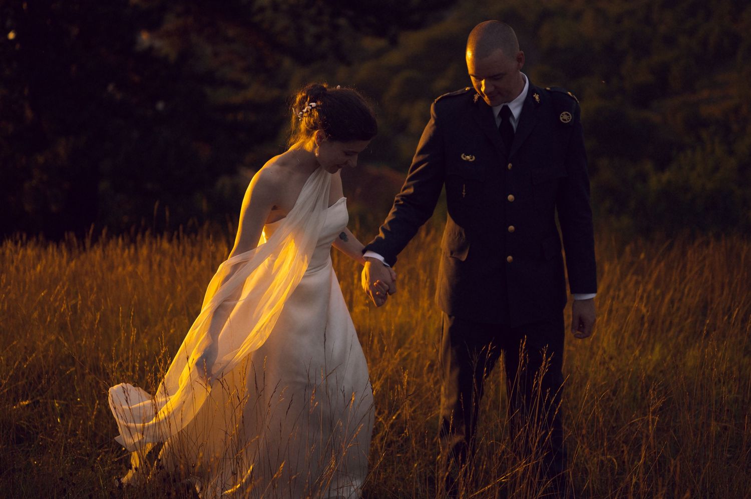 Military groom in dress uniform holds bride's hand walking through golden field at sunset during outdoor wedding.