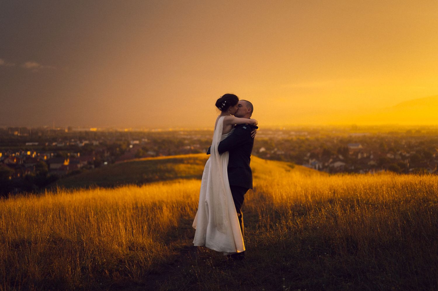 Couple embracing and kissing on a golden hill at sunset with a city skyline glowing in the background.