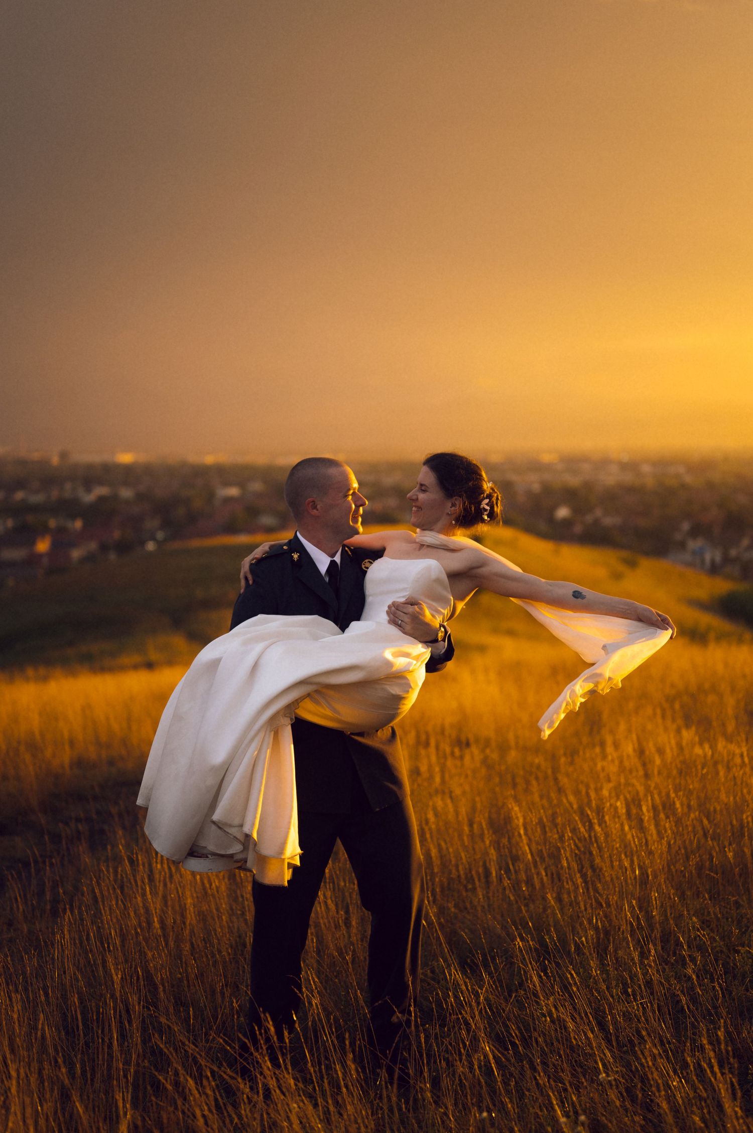 Groom carries bride in white dress through golden sunset field, city skyline glowing in warm light behind them.