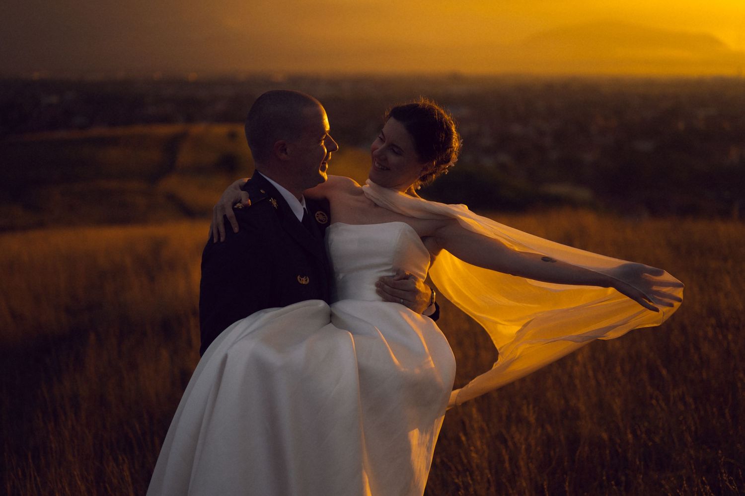 Bride and groom embrace at golden sunset in a wheat field, wedding dress veil flowing in the wind.