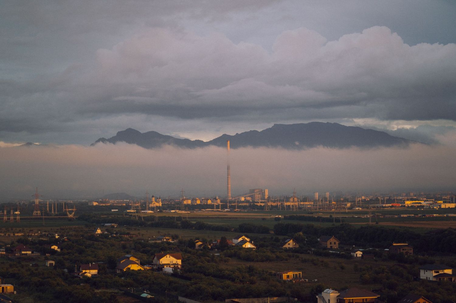 Industrial cityscape at dusk with dramatic storm clouds over mountains and a tall smokestack.