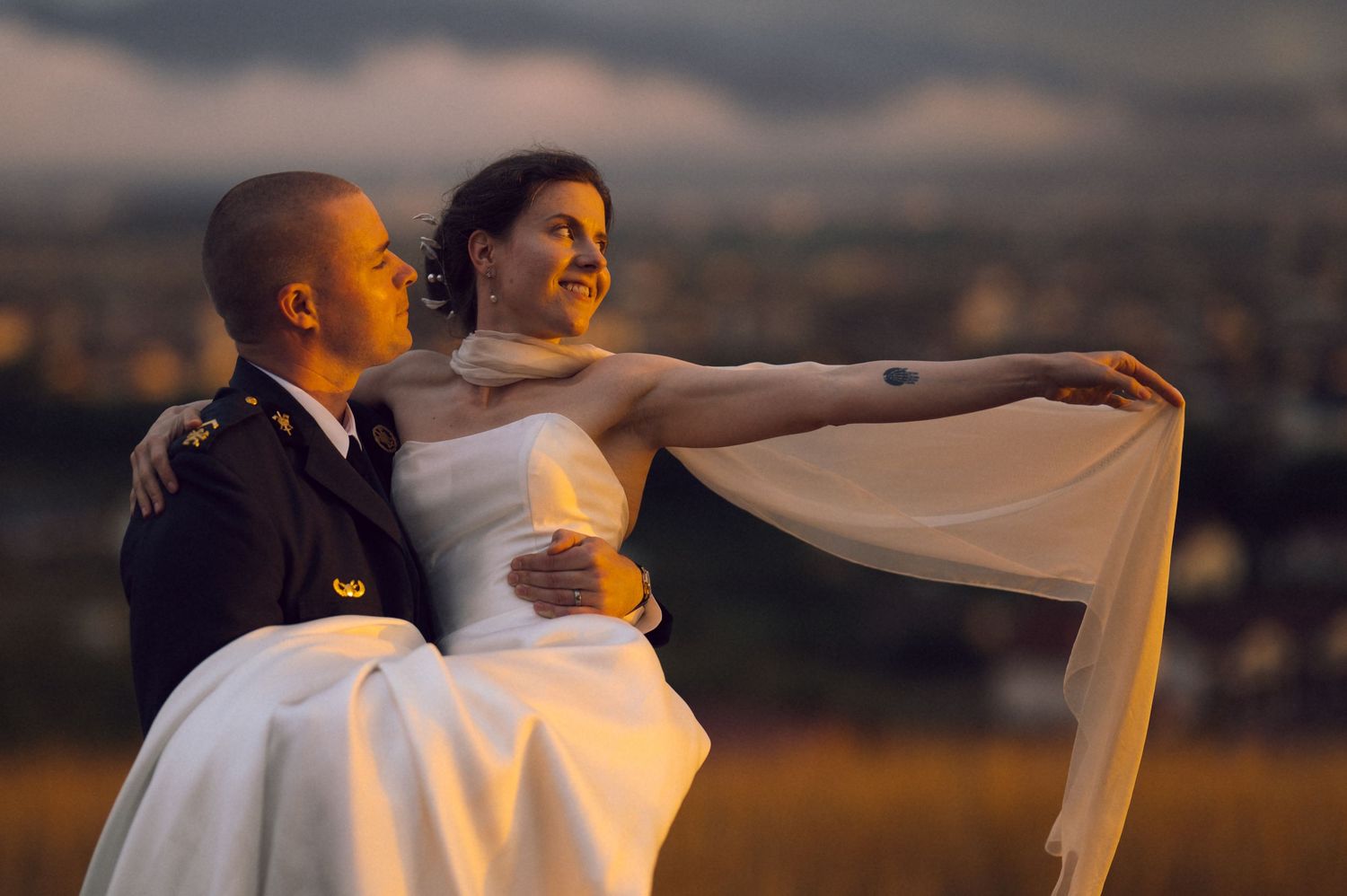 Groom in military uniform carrying bride in white wedding dress at sunset with city skyline backdrop.