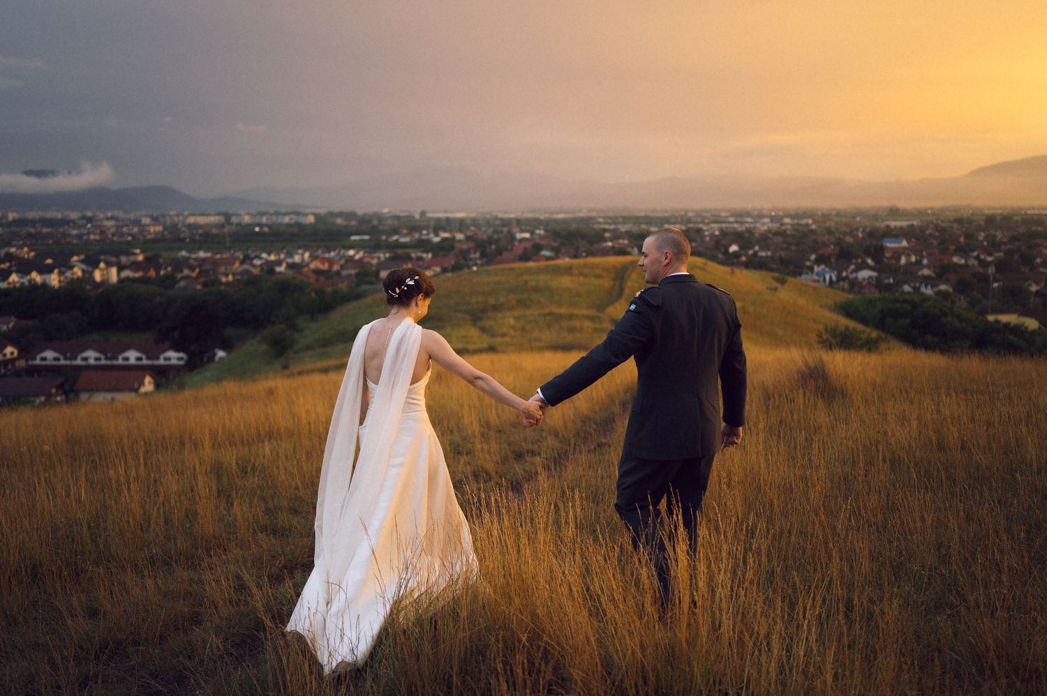 Bride and groom holding hands on a golden hill at sunset with a scenic cityscape and mountains in the background.