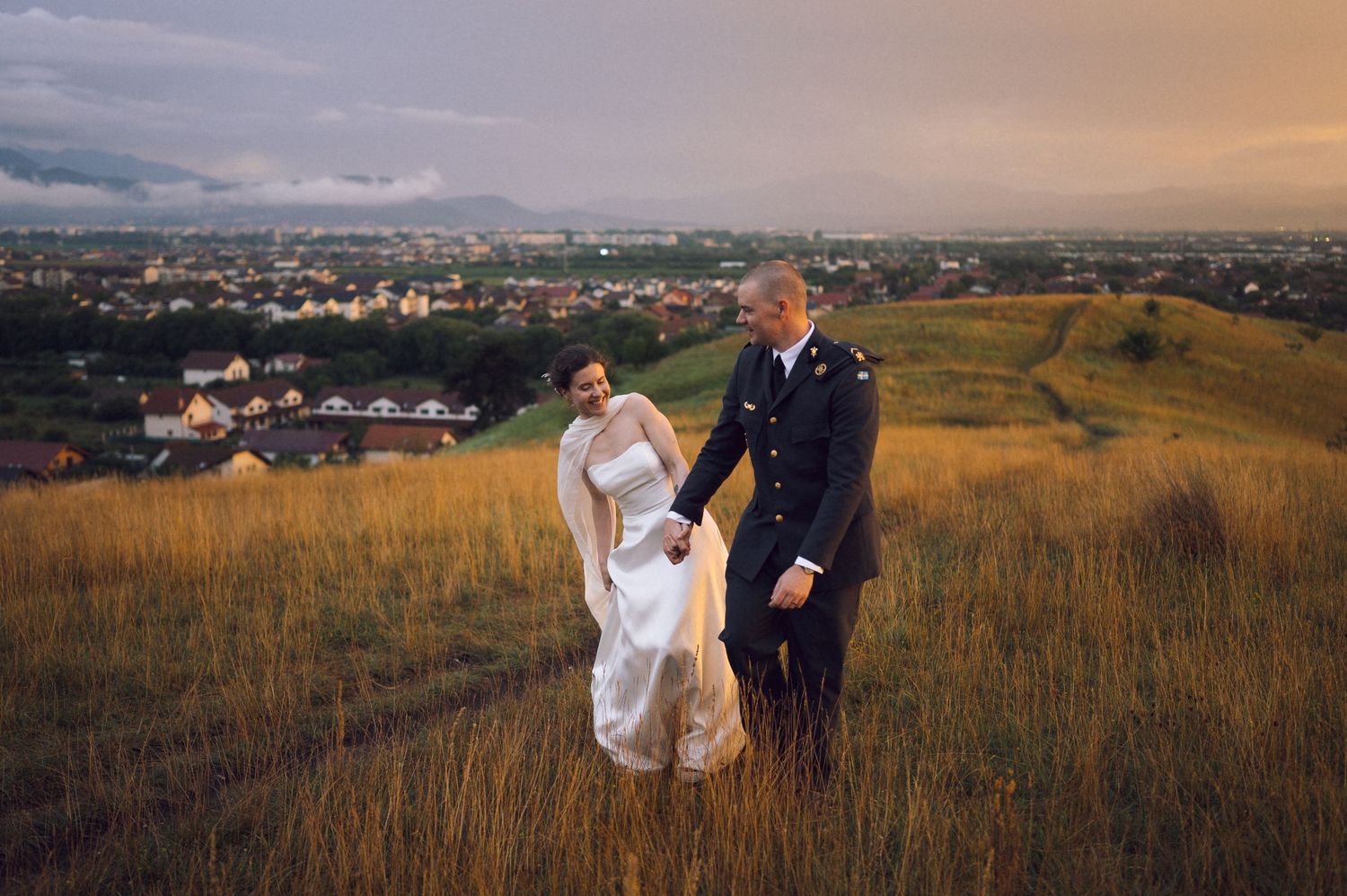 Bride and groom holding hands on a golden hillside with a scenic town and cloudy sky backdrop.