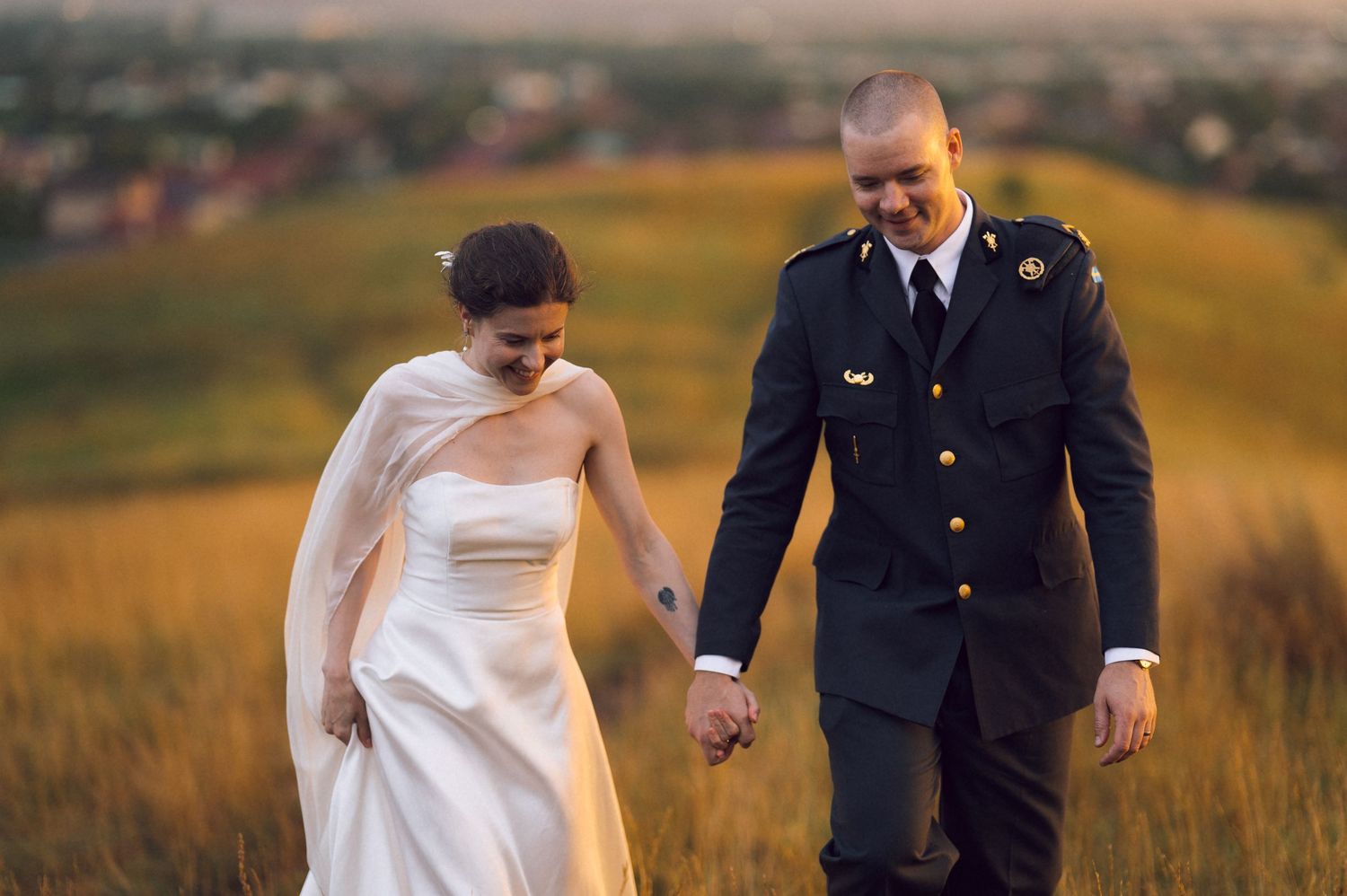 A bride in white gown and groom in military uniform walk hand in hand through a golden field at sunset.