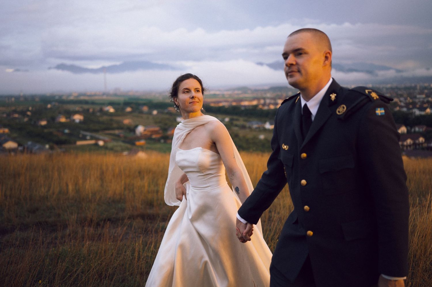 Military groom in uniform holds hands with bride in white dress on golden hillside with city skyline backdrop.