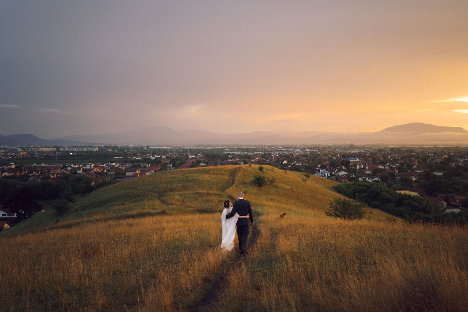 Bride and groom walking hand in hand on a grassy hilltop at sunset with a panoramic city view below.