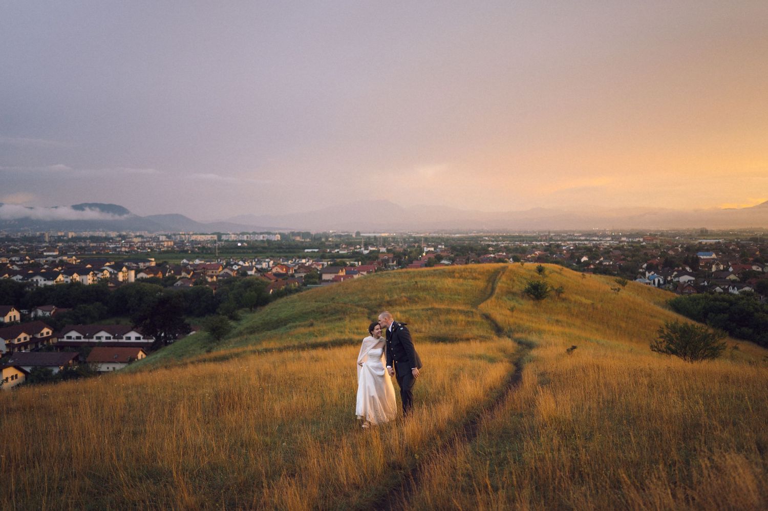 Couple in wedding attire embracing on golden hillside at sunset with panoramic city view below.