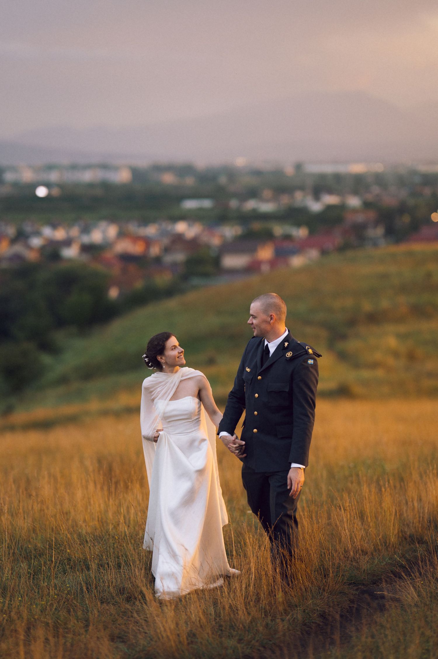 A bride in a white gown and groom in military uniform hold hands on a golden hillside at sunset with a town below.