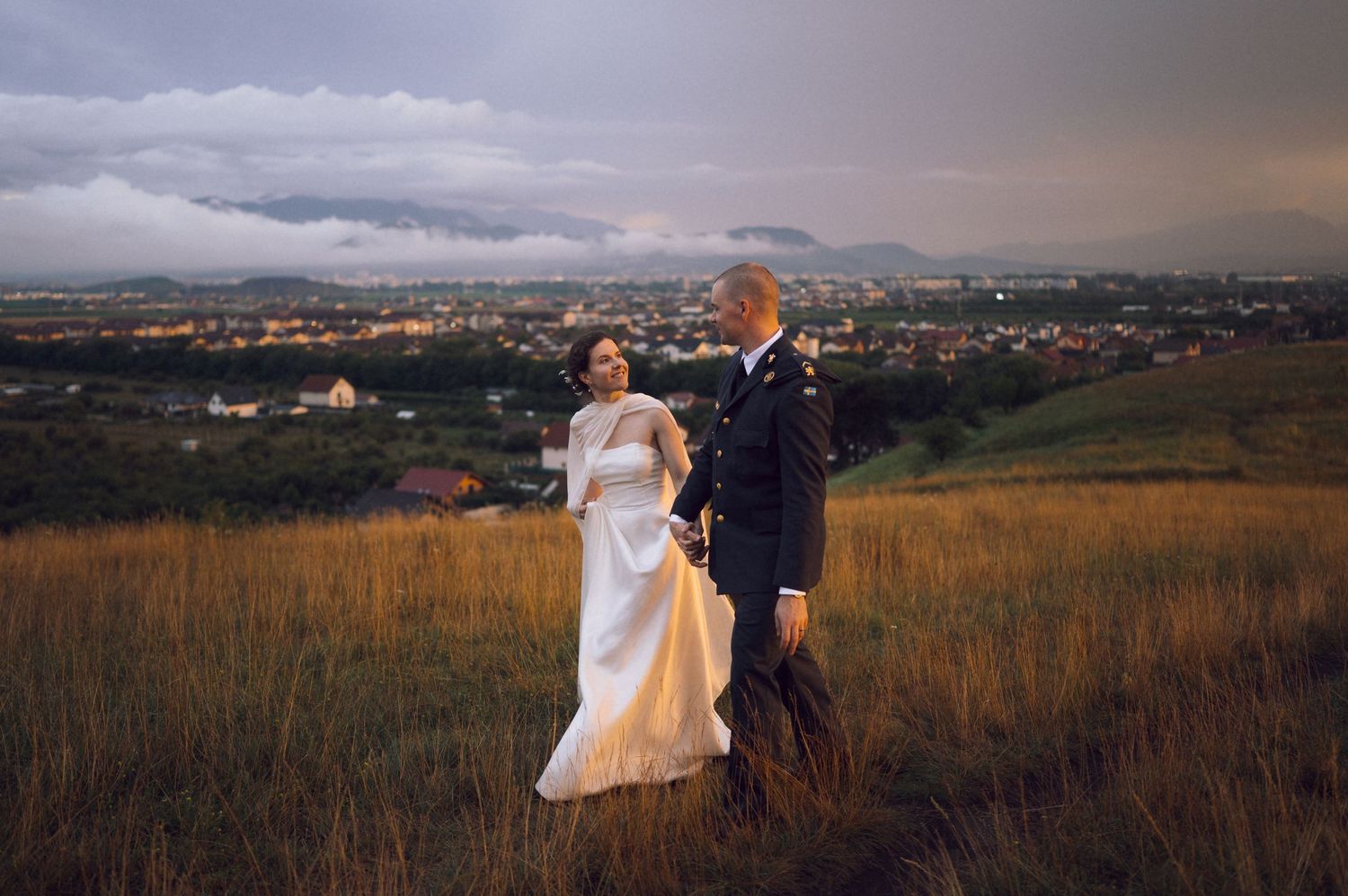 Bride and groom walking on a grassy hill at dusk with a scenic city skyline in the background.