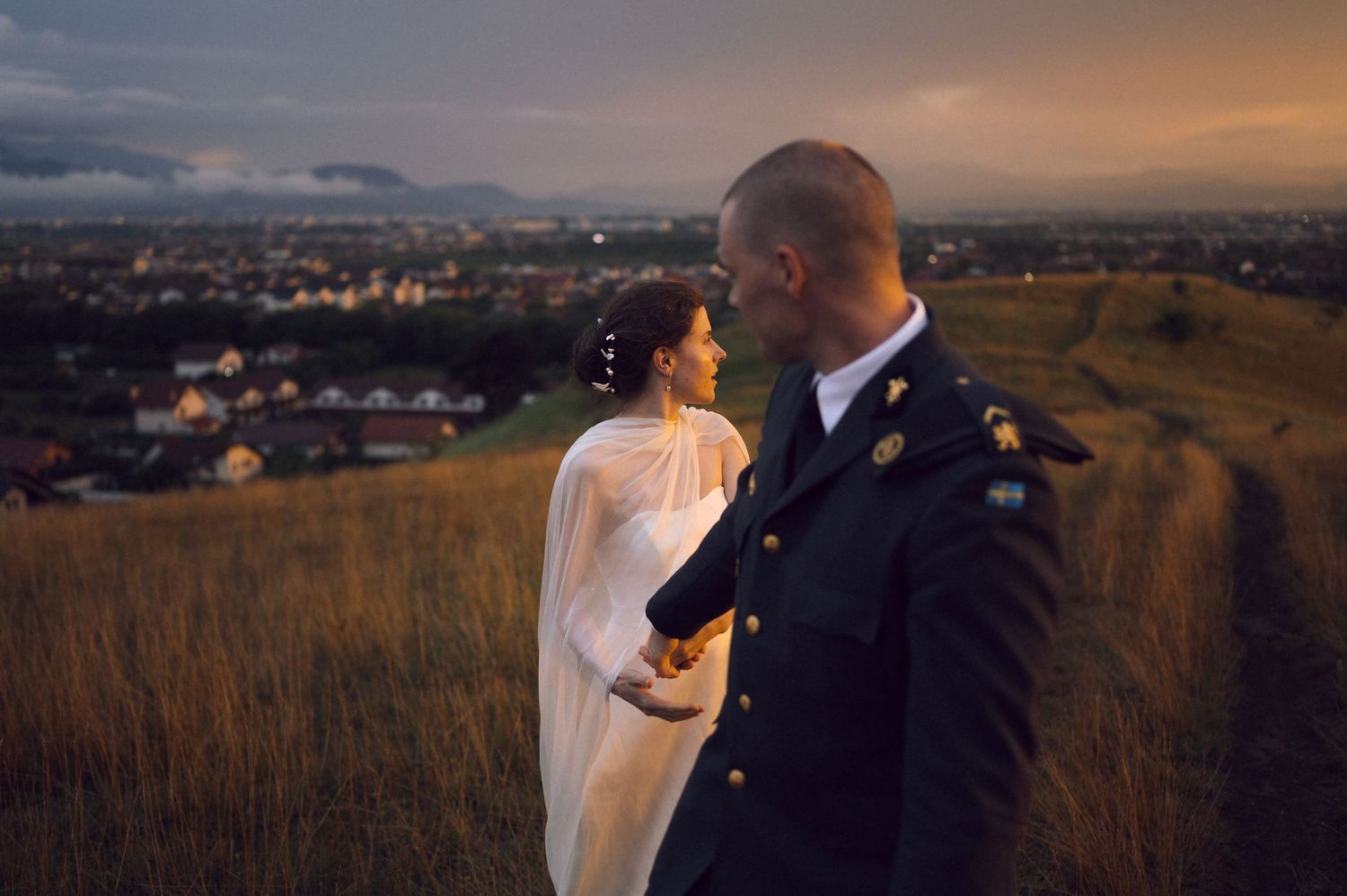 Military groom and bride in white cape share romantic moment on hilltop at dusk with city lights below.