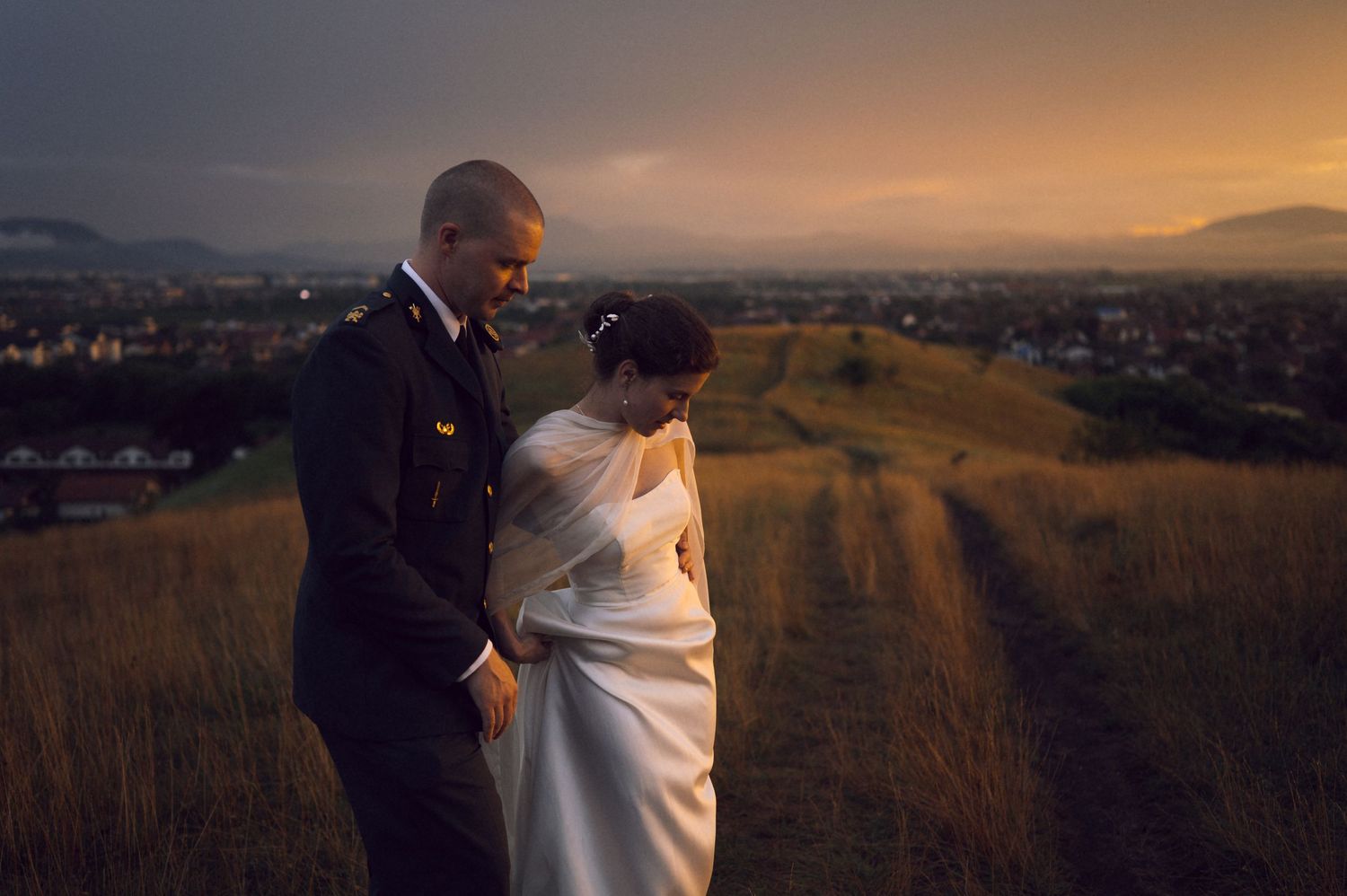 Bride and groom share a romantic moment on a golden hill at sunset with a scenic cityscape and mountains in the background.