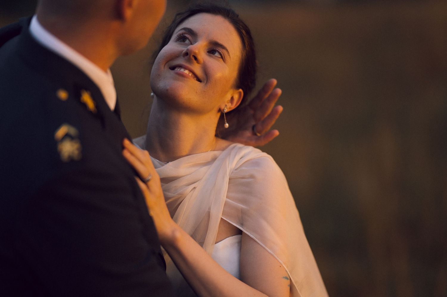 Bride in white dress smiling up at groom in military uniform during romantic outdoor wedding ceremony.