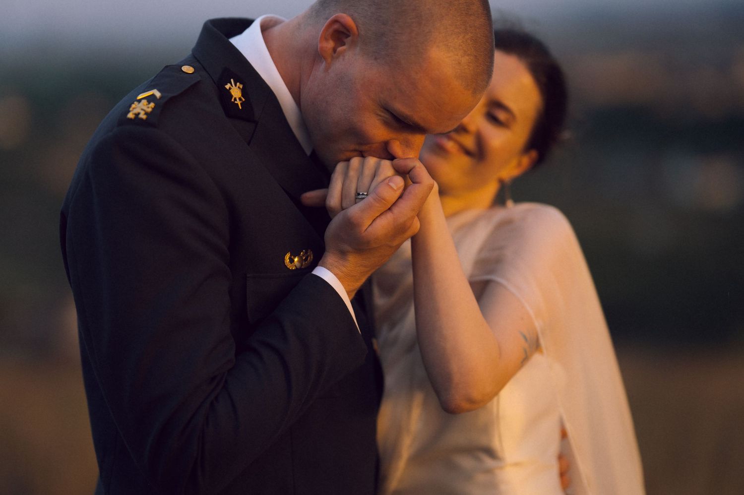 Military groom kissing bride's hand during romantic outdoor wedding ceremony at sunset.