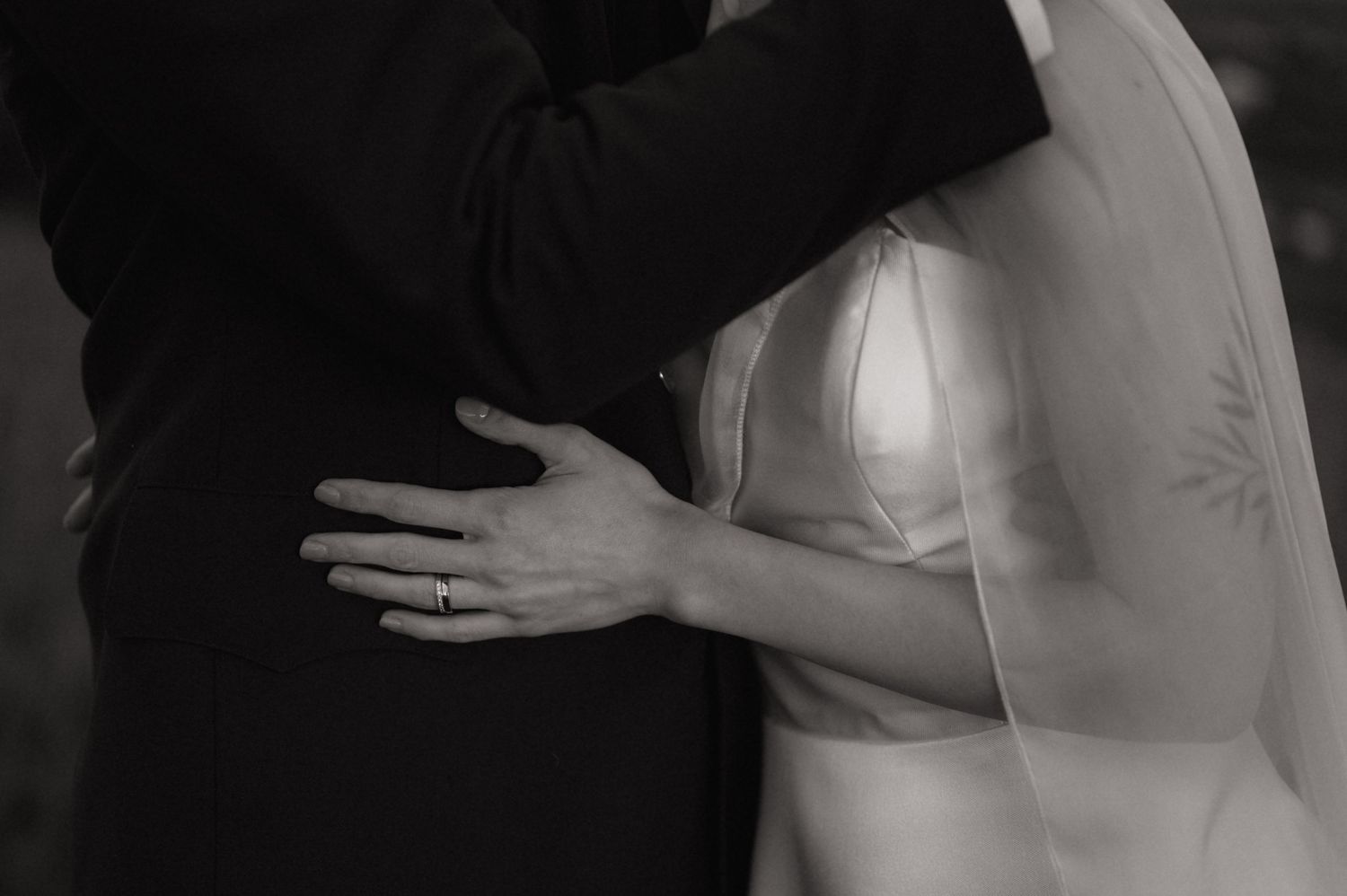 Black and white photo of bride and groom embracing, showing wedding ring on brides hand and elegant veil.