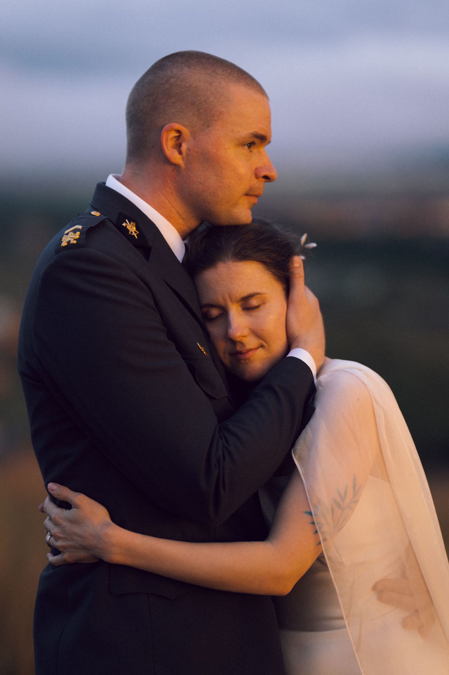 Military groom in uniform embraces bride in white veil during romantic outdoor wedding portrait at sunset.