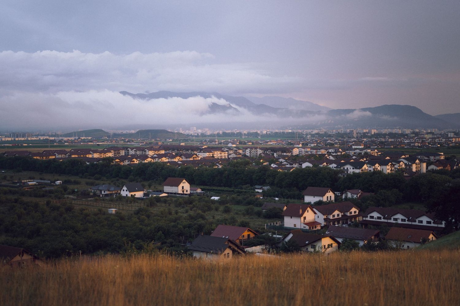 Aerial view of a suburban town nestled below misty mountains at dusk with golden grasslands in foreground.