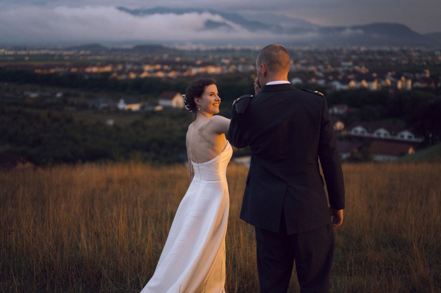 Bride and groom standing on a golden hillside at sunset with a panoramic city view and dramatic cloudy sky.