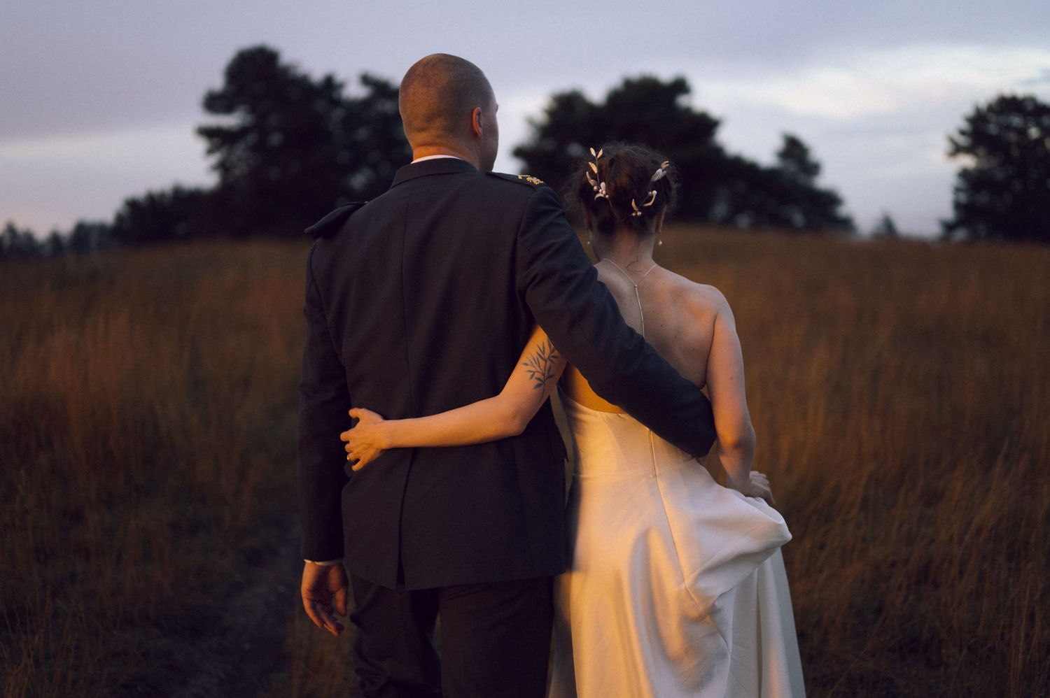 A bride and groom walk hand in hand through a golden field at dusk, viewed from behind.