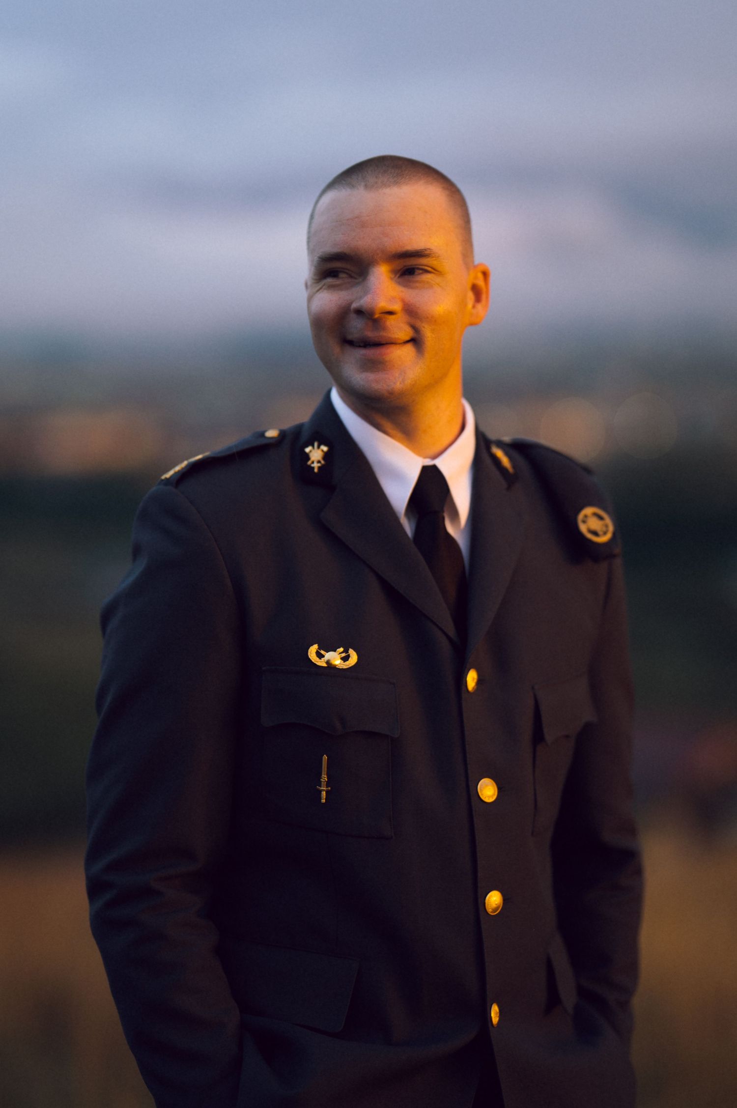 Young man in dark military dress uniform with gold buttons smiling outdoors at dusk.