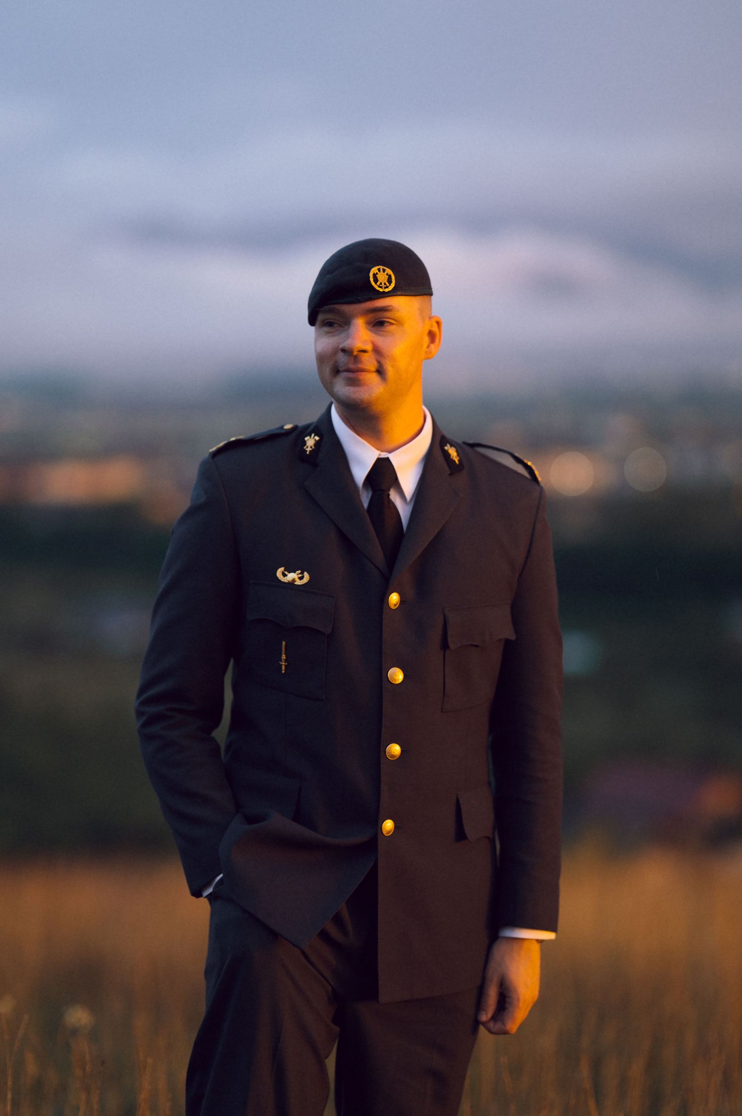 Military officer in dark dress uniform and black beret standing in a field at golden hour.