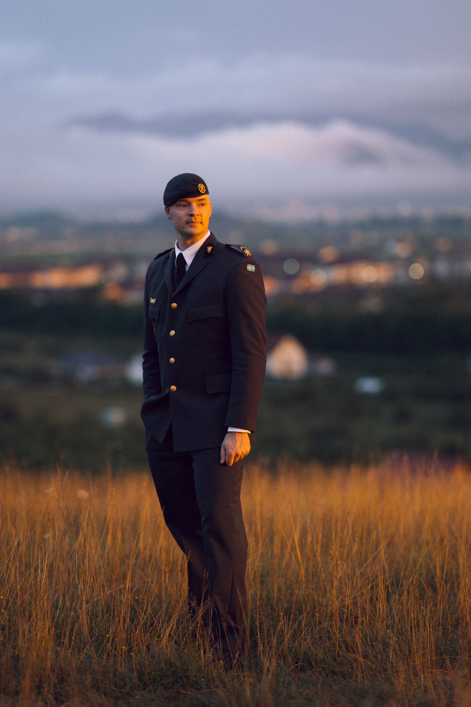 Young man in dark military dress uniform and beret standing in golden field at dusk with city lights behind.