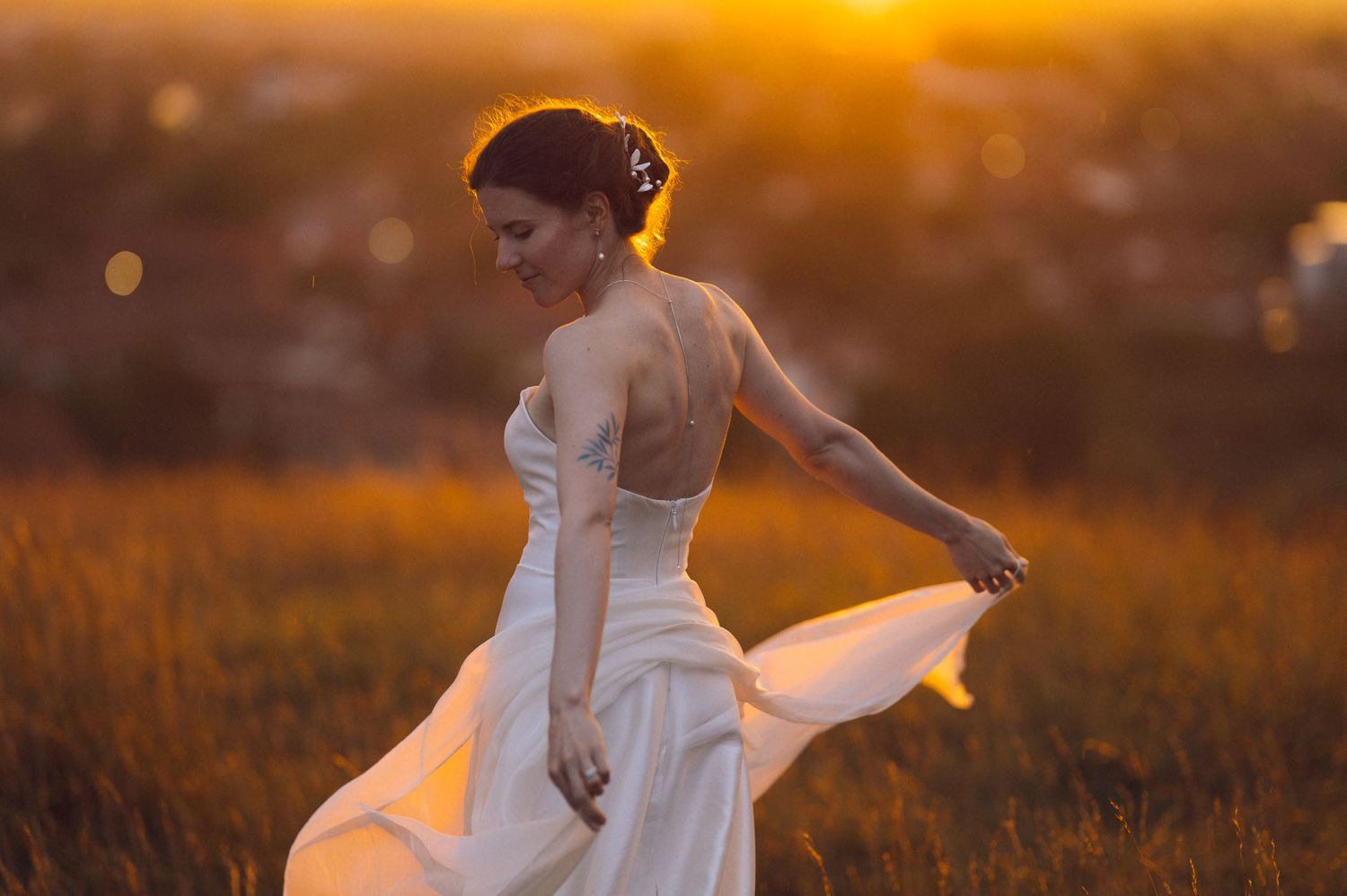 Bride in white backless gown twirling in golden sunset field, hair up with floral accessories.