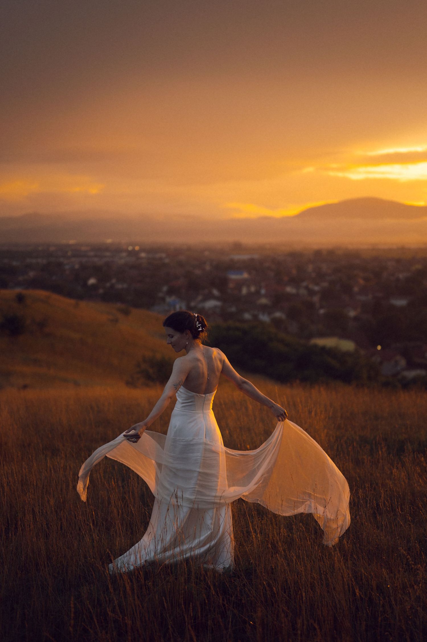 Bride in white flowing gown twirling in golden field at sunset with mountain cityscape backdrop.