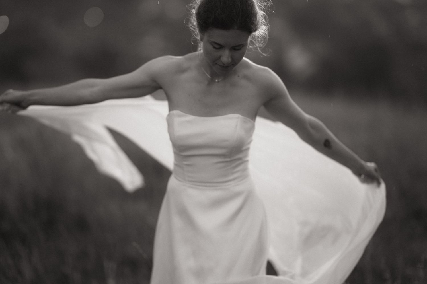 Black and white photo of a bride in a strapless gown twirling her dress in an outdoor field.