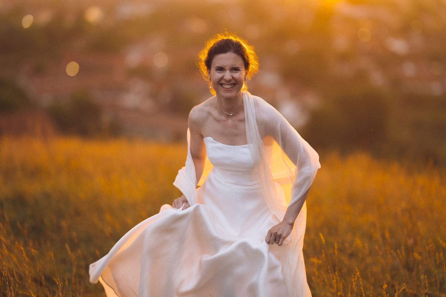 Smiling bride in white gown running through golden sunlit field at sunset, wearing floral hair wreath.