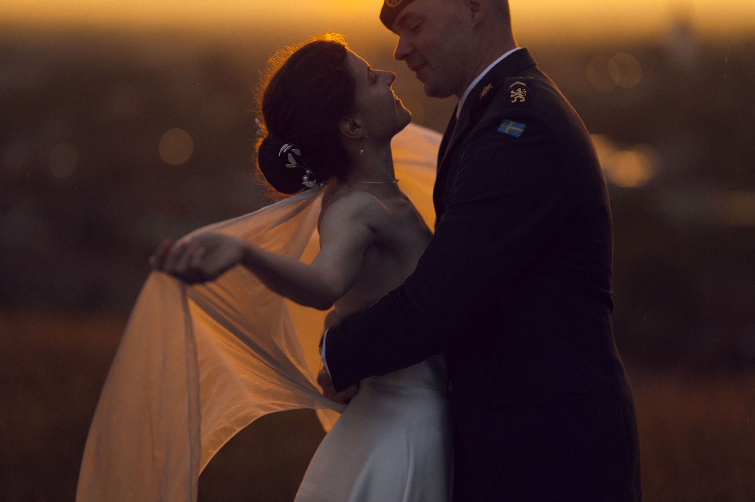 Couple sharing a romantic embrace at sunset, bride in white gown with flowing veil, groom in military dress uniform.