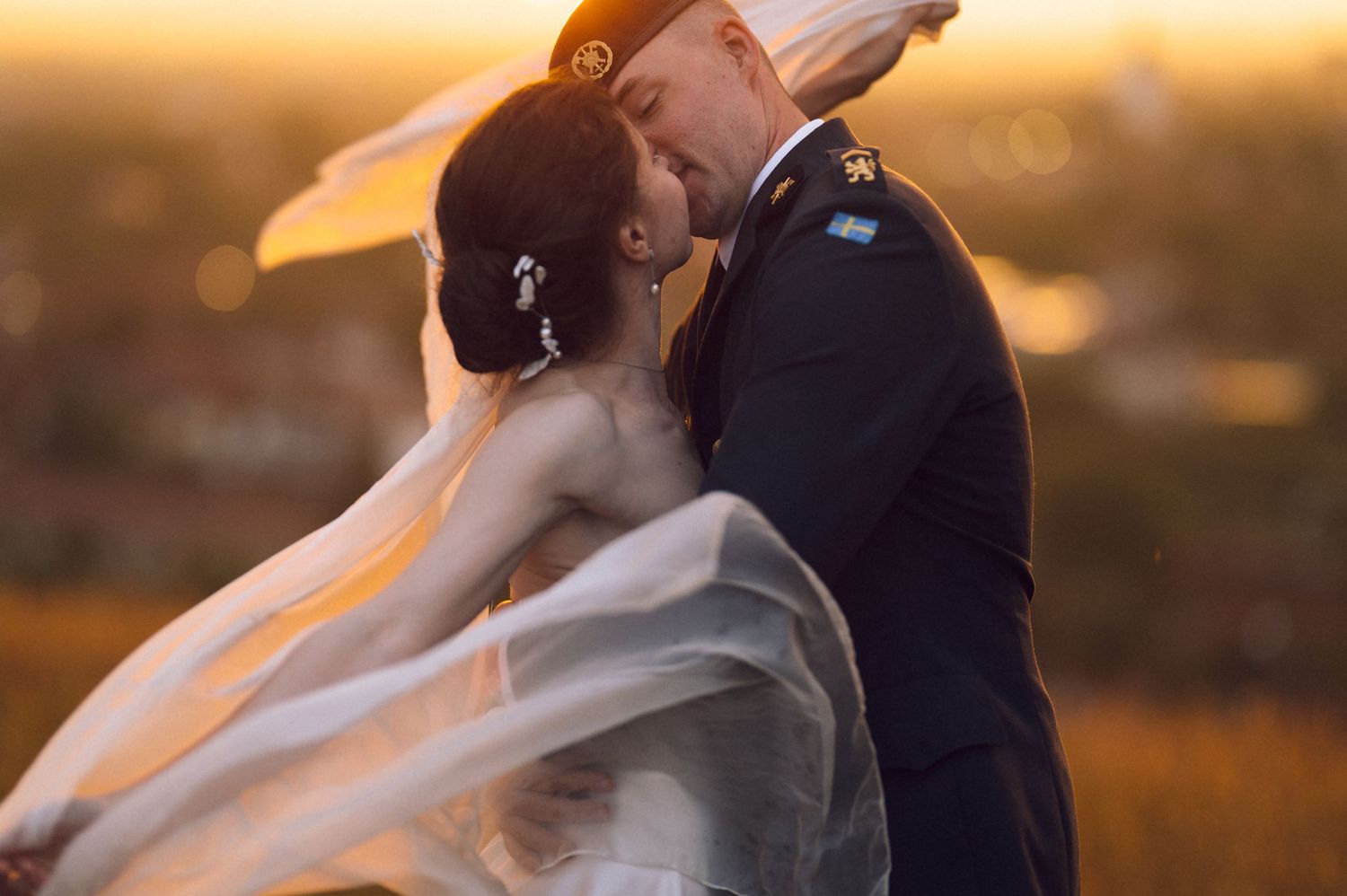 A couple shares a romantic kiss at sunset, bride's veil flowing in the wind, groom in military dress uniform.