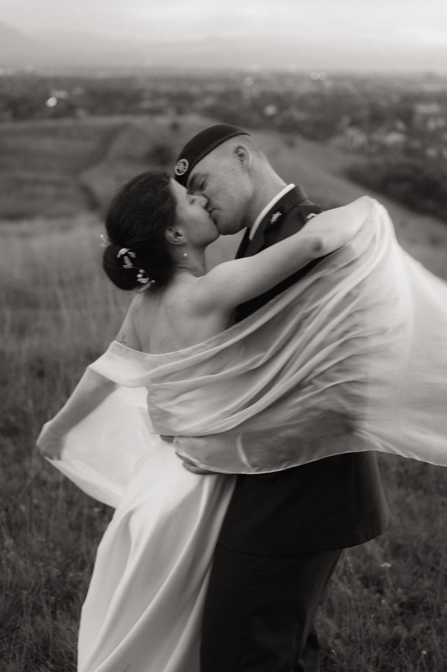 Black and white photo of a bride and groom kissing in a field, her veil flowing in the wind.