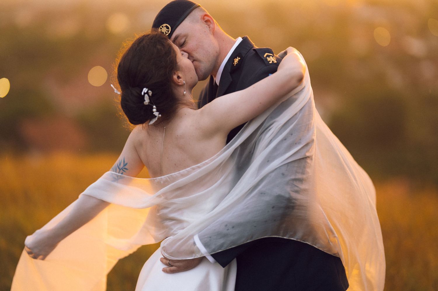 Military groom in uniform lifts bride in white gown and veil, sharing romantic kiss at golden hour outdoor wedding.