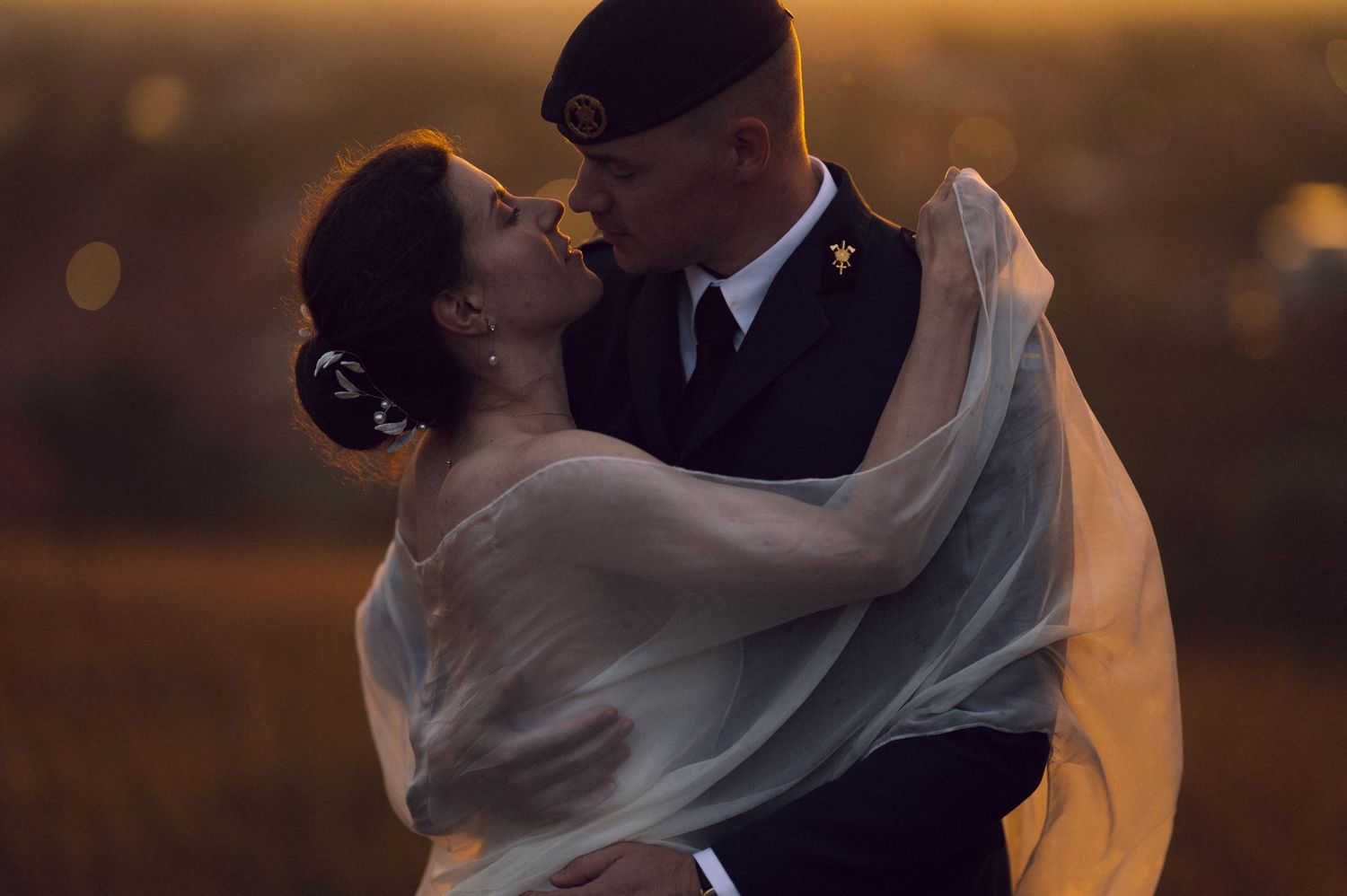 Military groom in uniform kissing bride in white veil at golden sunset in romantic outdoor wedding portrait.