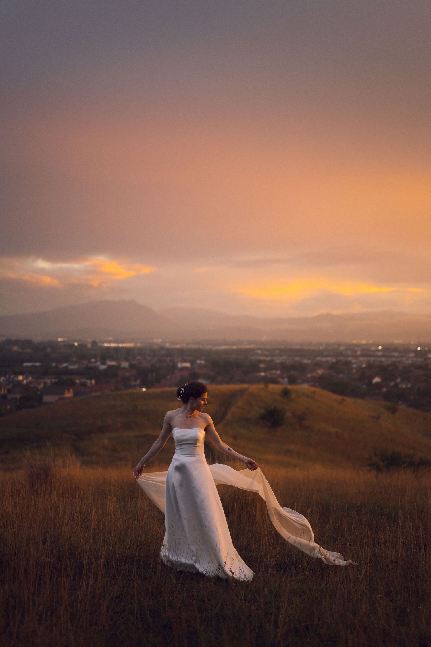 Bride in white gown with flowing veil stands on golden hilltop at sunset with city and mountains backdrop.