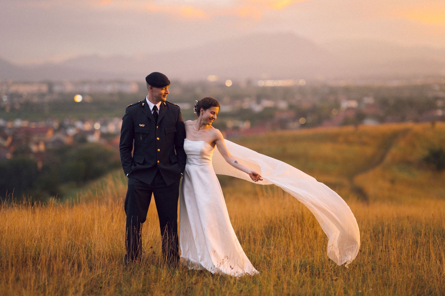 Couple in wedding attire standing in golden field at sunset, man in military uniform, woman in flowing white gown.
