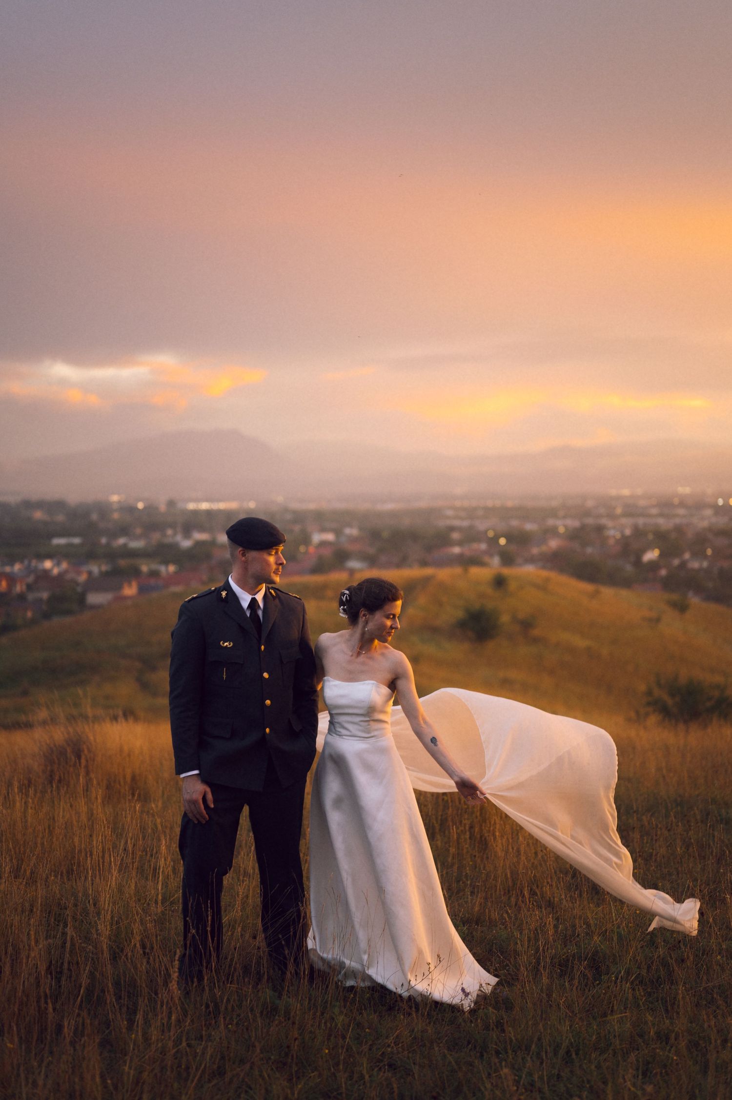 Couple in military uniform and wedding dress posing on a grassy hill at sunset with mountains in the background.