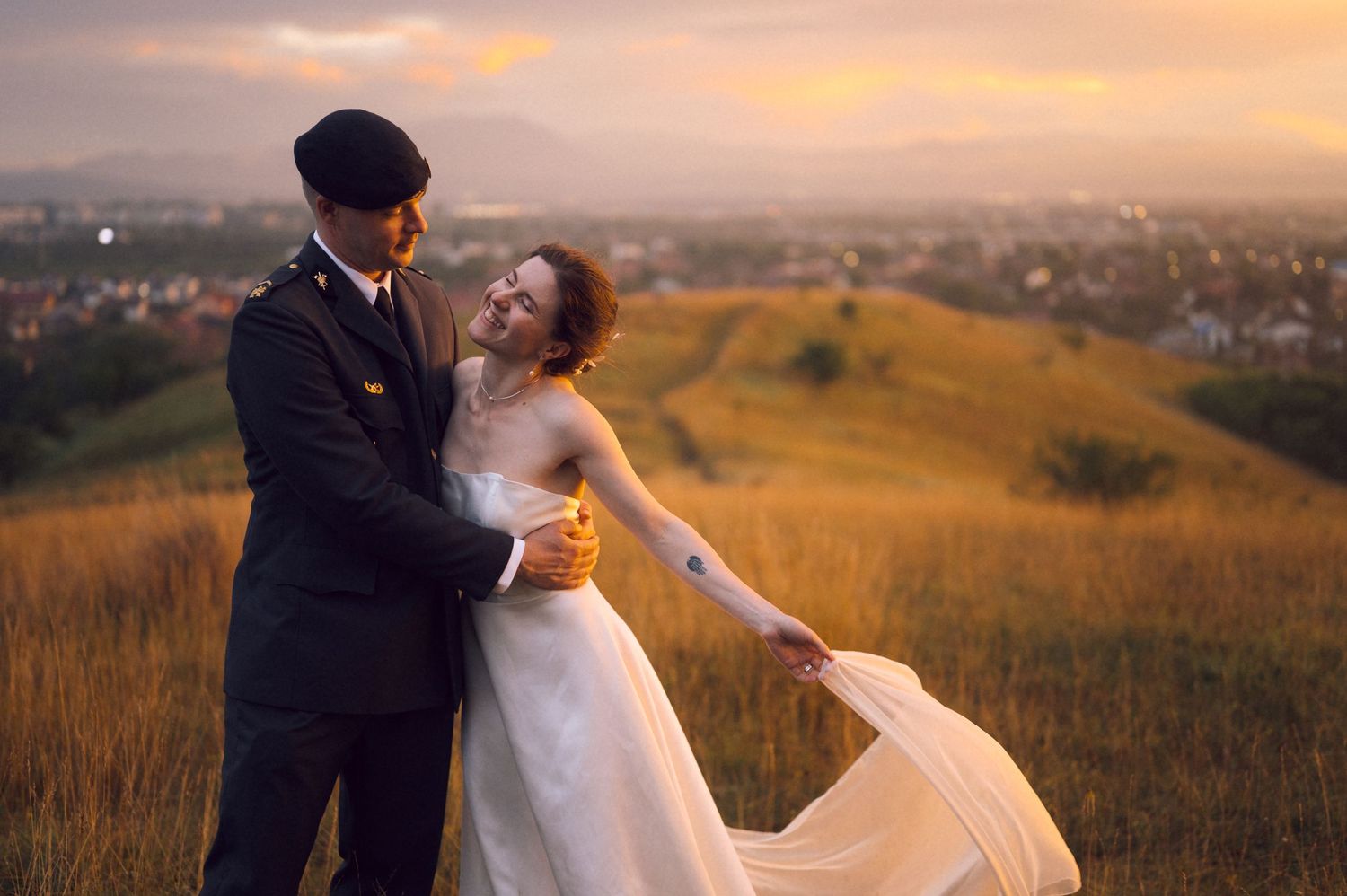 Couple in wedding attire embracing on a golden hillside at sunset with a scenic cityscape in the background.