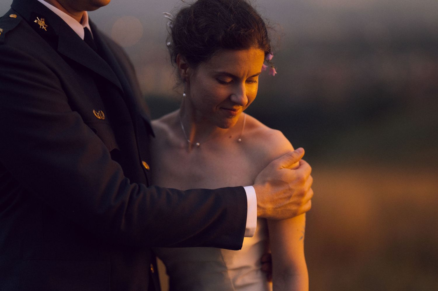 A groom in military uniform tenderly holds a bride's shoulder during a golden hour outdoor wedding portrait.