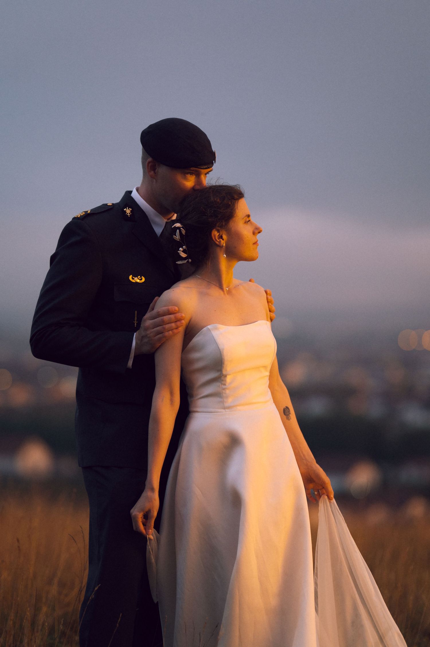 Military groom in black uniform embraces bride in white gown at sunset with city lights in background.