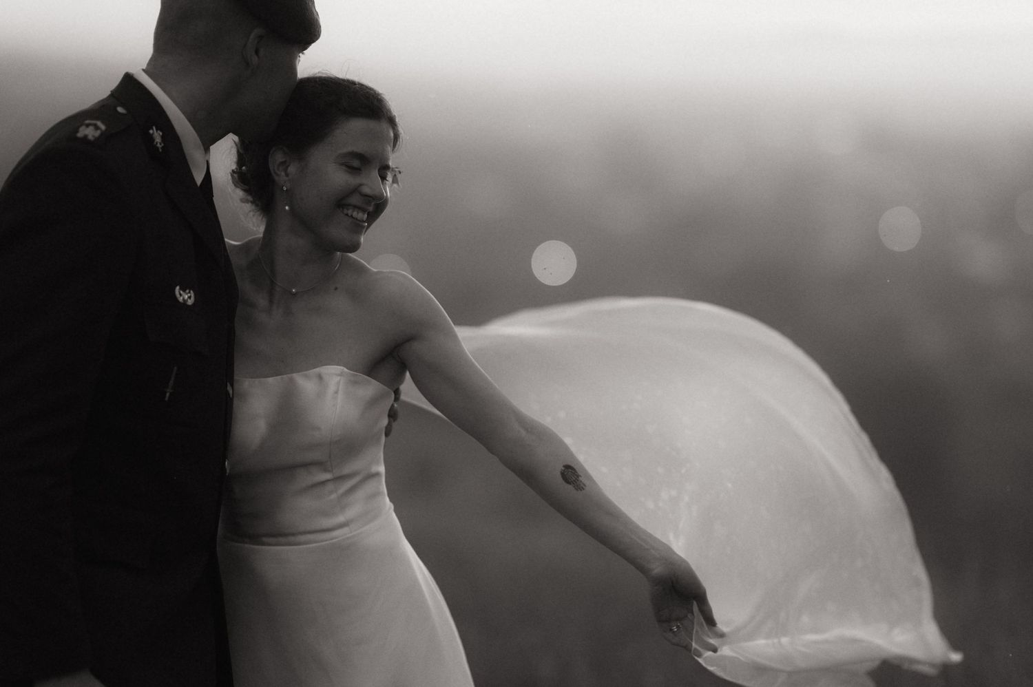 Black and white photo of a smiling bride in a strapless gown with veil blowing in wind beside a groom in military uniform.