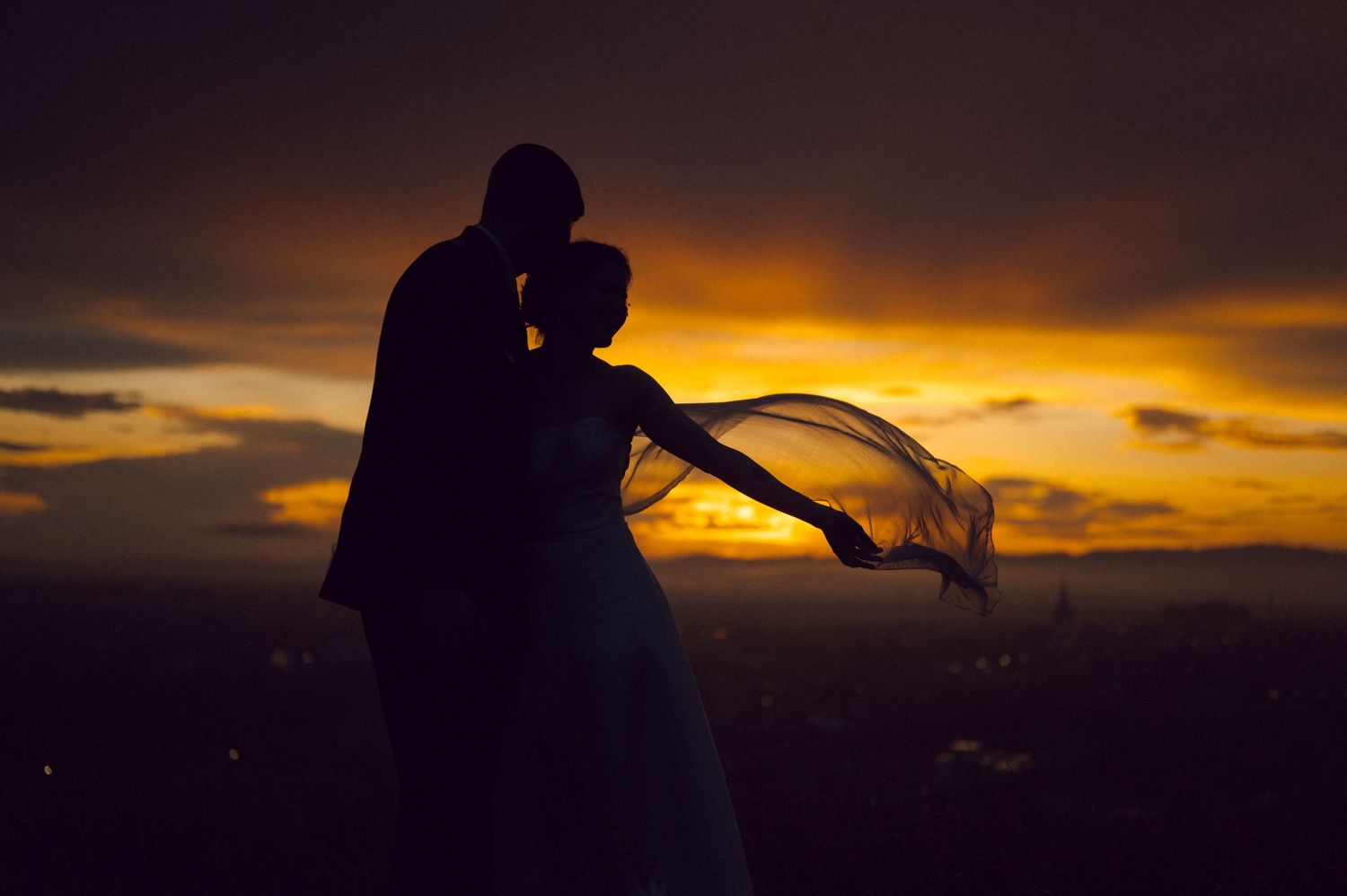 Silhouette of a couple embracing at sunset with a flowing wedding veil, romantic golden sky backdrop.
