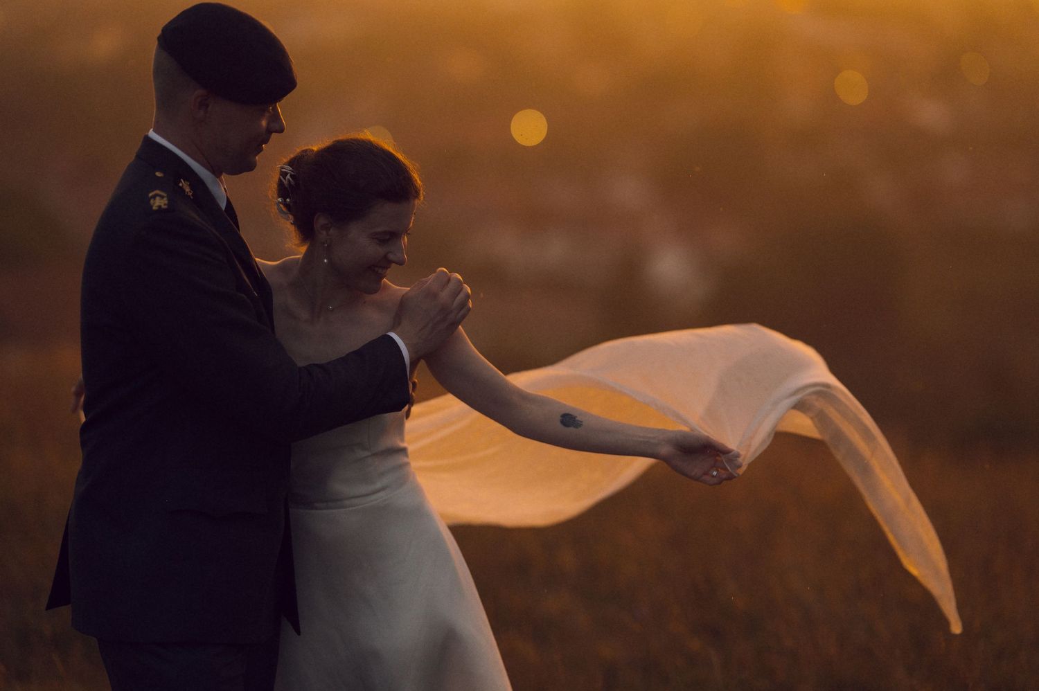 Couple dancing in golden sunset light in a field, bride in white gown with veil flowing, groom in military uniform.