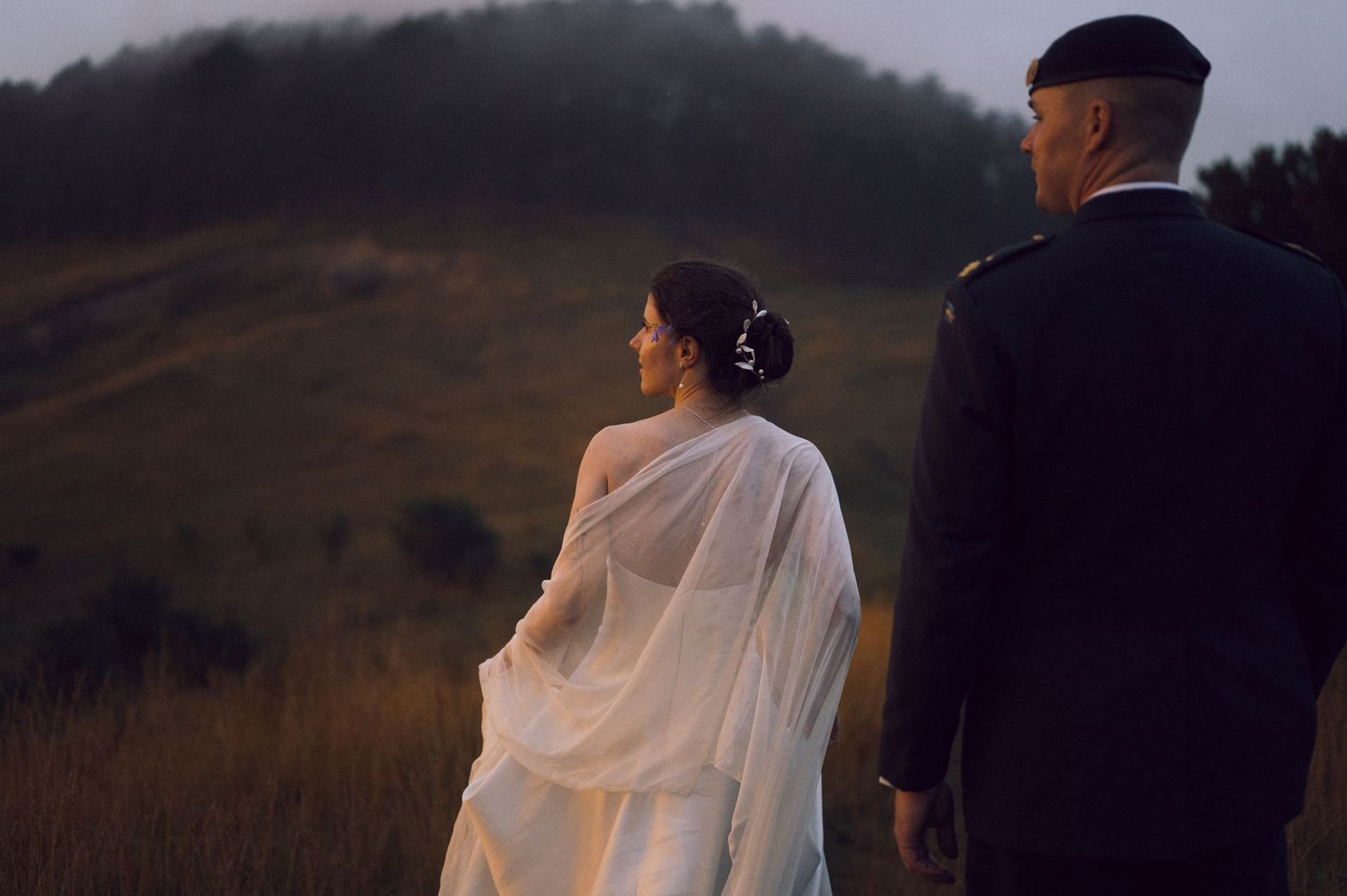A bride in a white flowing gown and groom in military uniform walk through a misty green field at dusk.