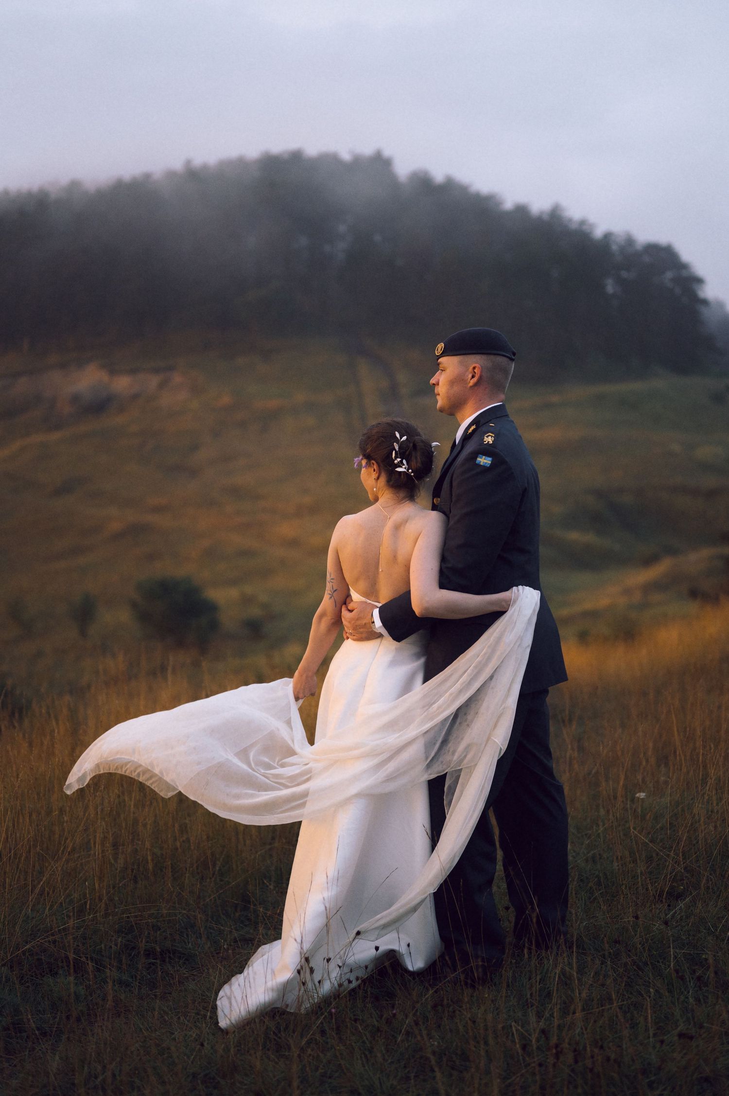 Military groom in uniform embraces bride in flowing white dress, standing in misty green field at dusk.