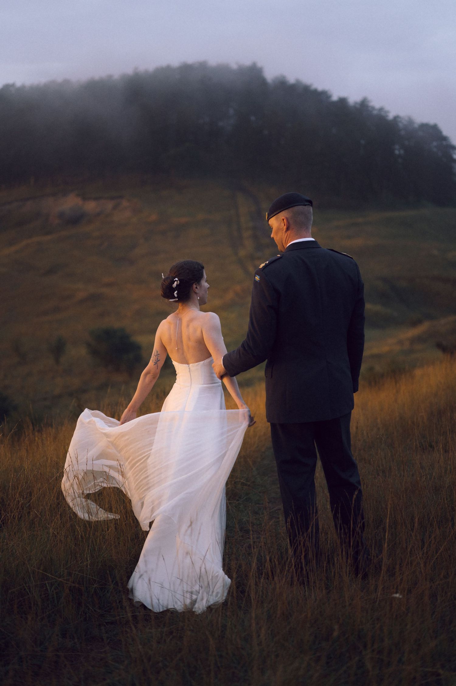 A bride in a flowing white backless gown holds hands with a groom in military uniform walking through misty hills.