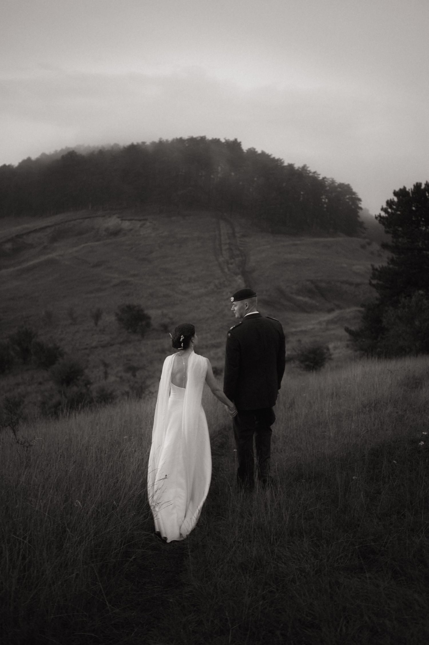Black and white photo of a bride and groom walking hand in hand through a misty hillside meadow.